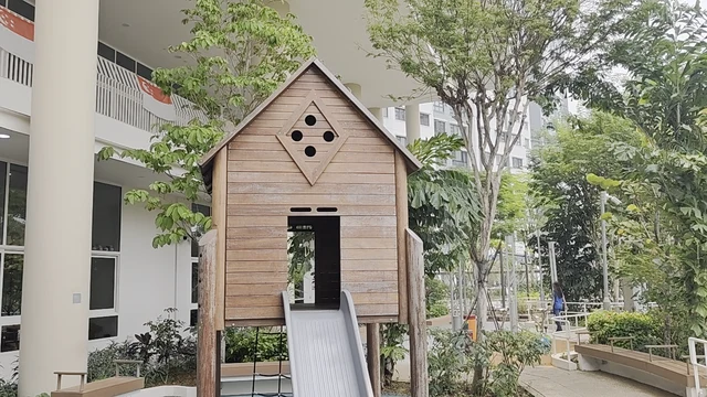 An outdoor play area features a wooden playhouse with a grey slide, situated on a paved surface surrounded by lush greenery and trees. a white building with large windows and a balcony is visible, while a wooden bench and white railings line a pathway. The scene is bathed in natural daylight, creating a serene and inviting atmosphere for children to play.