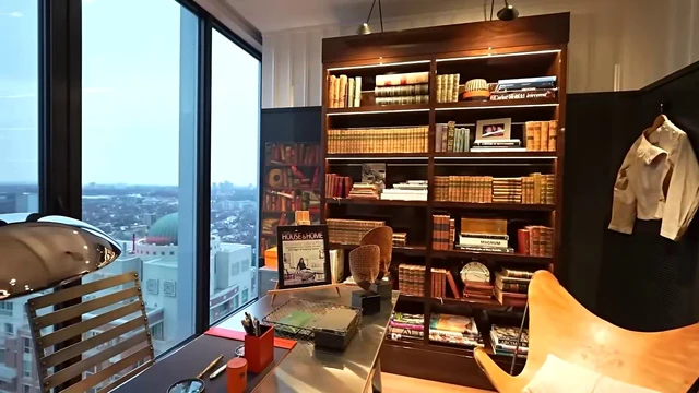 A modern home office scene featuring a large window with a view of a city skyline and a domed building. A dark wood bookshelf filled with books and decorative items stands against a wall, illuminated by warm lighting. A desk with a lamp, stationery, and a magazine is positioned near the window, accompanied by a wooden chair. A colorful framed artwork hangs on the wall, adding a vibrant touch to the room. The atmosphere is cozy and intellectual, with a focus on design and functionality.