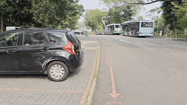 A scene featuring a black hatchback parked on a paved surface composed of interlocking bricks, positioned adjacent to a red-painted curb. The vehicle displays visible scuff marks on its rear bumper and wheel arch. a wide asphalt road extends into the distance, marked with a faded red directional arrow painted on the ground. two large buses are visible on the road, one light blue and one green and white. The environment is lined with dense green trees and vegetation, suggesting a park or campus setting. The lighting is bright and diffuse, indicating daytime conditions.