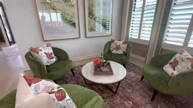 A bright, airy interior scene features a cozy seating arrangement centered around a round, white marble coffee table with a dark metal base. The table rests on a patterned rug and holds a wooden tray with a vase of orange flowers, a bottle, and small metallic cups. Three plush green armchairs with a subtle geometric texture surround the table, each accented with a floral pillow featuring pink and red blooms. The room is illuminated by natural light streaming through large windows covered with white plantation shutters. Two large framed artworks hang on the white walls, and a glass bar cart is visible in the corner, contributing to the room's elegant and inviting atmosphere.