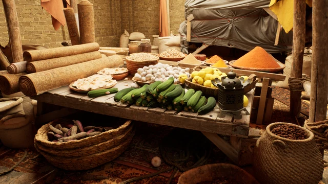 A rustic market stall is set up within a structure featuring exposed brick walls and wooden support beams. A low wooden table serves as the central display, laden with fresh produce including a large pile of green zucchinis, a bowl of lemons, and a plate of flatbread. Surrounding the table are woven baskets containing dried goods and a large sack filled with brown beans. Rolled carpets lean against the back wall, while various piles of colorful spices, including a prominent mound of orange powder, are arranged on the table and nearby surfaces. The scene is illuminated by warm, ambient lighting that highlights the textures of the wood, fabric, and food items.