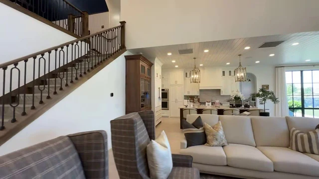 A spacious, open-concept interior features a grand staircase with dark wood railings and iron balusters ascending to a second-floor landing with a dark ceiling. The living area is furnished with a large beige sofa, two grey plaid wingback armchairs, and a round white coffee table holding a vase of greenery. A tall wooden cabinet stands near the stairs, while the background reveals a kitchen with white cabinetry, stainless steel appliances, and pendant lighting. Large windows and glass doors allow natural light to illuminate the room, which is decorated with various throw pillows and framed artwork on the walls.