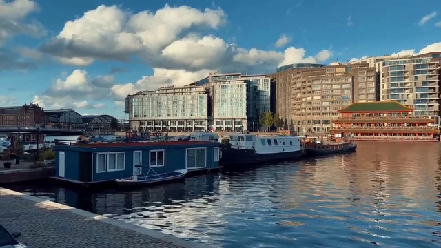 A canal scene featuring a row of moored houseboats, including a blue vessel with a small dinghy alongside and a white boat further down the line. The water reflects the bright blue sky and scattered white clouds. The far bank is lined with a mix of modern glass-fronted buildings and traditional brick architecture, including a distinctive structure with a green roof. A paved quay runs along the near side, where a white car is parked. The lighting is bright and sunny, casting distinct shadows and highlighting the textures of the buildings and the ripples on the water surface.