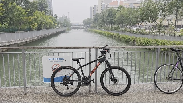 A canal scene featuring a paved walkway bordered by a metal railing. Two bicycles, a black mountain bike and a black and purple road bike, are parked against the railing. A white sign with blue text reading 'NO FISHING' is attached to the railing. The canal water is calm and green, flanked by concrete embankments and dense vegetation. In the distance, residential buildings and trees are visible under an overcast sky.